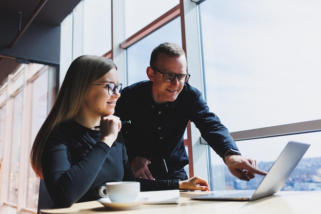 Smiling young male and female manager sitting at table looking at computer screen explaining enterprise software or working on online project with colleagues