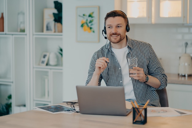 Smiling young male freelancer in headset taking part in online meeting or web conference on laptop, holding glass of water and pen while working at home. Remote job concept