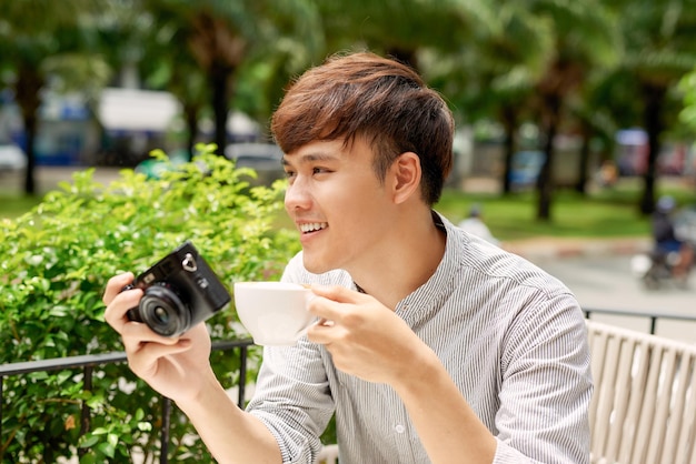 Smiling young male sitting at table in cafe with photo camera while drinking cup of coffee