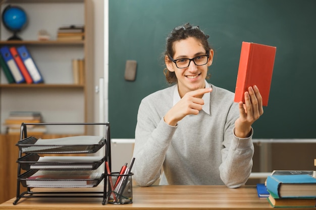 Photo smiling young male teacher holding and points at book sitting at desk with school tools on in classroom