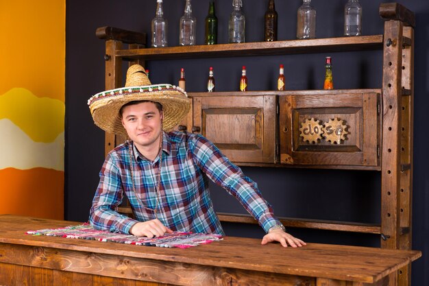 Smiling young man as bartender in a sombrero leaned on bar counter in Mexican pub, a shelf with spicy sauces and empty bottles in the background