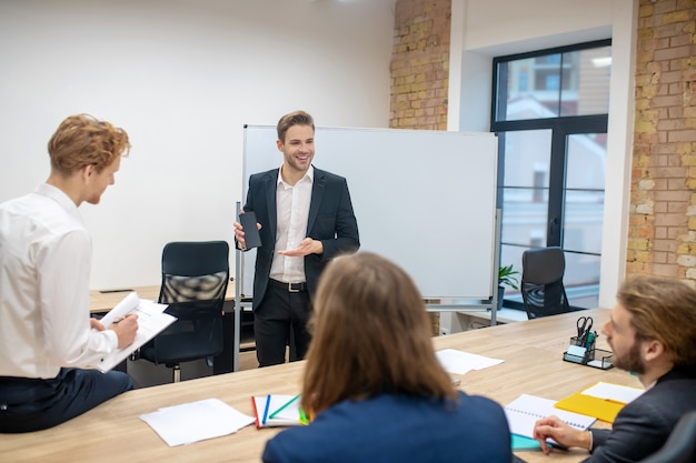 Smiling young man in business suit standing at stand showing smartphone and sitting interested colleagues