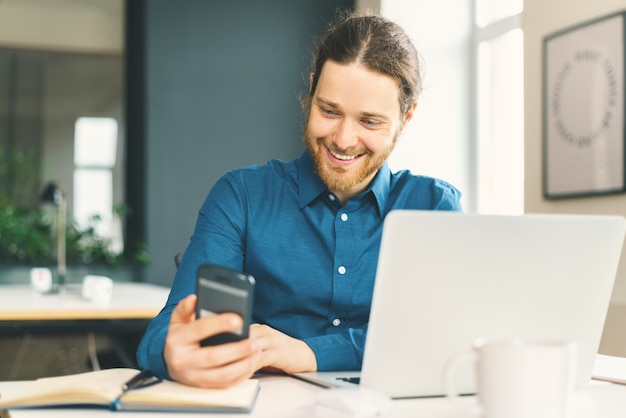 Smiling young man in casual wear looking at smartphone screen at workplace