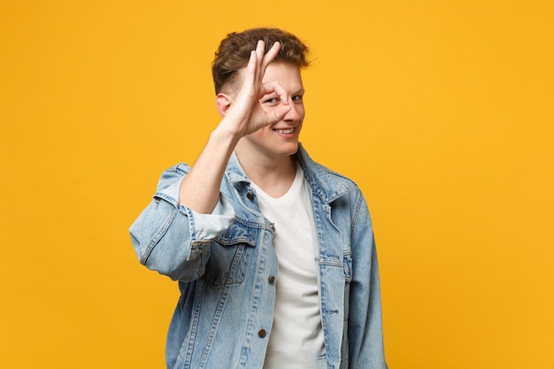 Smiling young man in denim casual clothes holding hand near eyes, imitating glasses or binoculars isolated on yellow orange background. People sincere emotions, lifestyle concept. Mock up copy space.