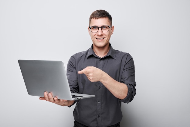 smiling young man in glasses holding laptop and pointing to screen on gray background