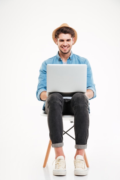 Smiling young man in hat sitting on chair and using laptop