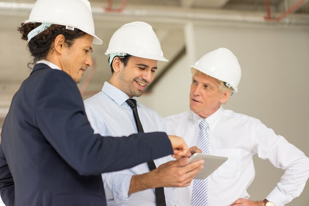 Smiling young man in helmet and tie showing something to colleagues on tablet screen 