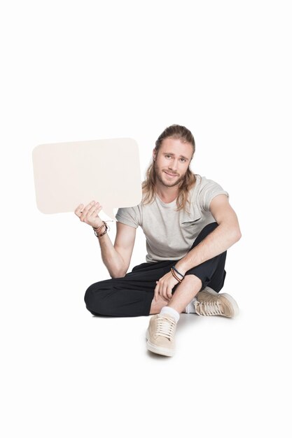 Smiling young man holding empty speech bubble and looking at camera isolated on white