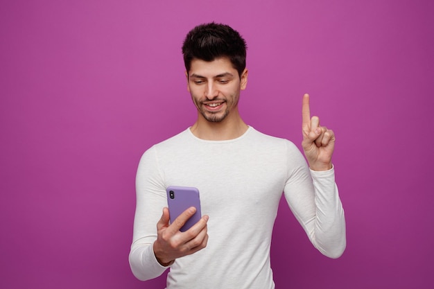 Smiling young man holding and looking at mobile phone poinitng up isolated on purple background