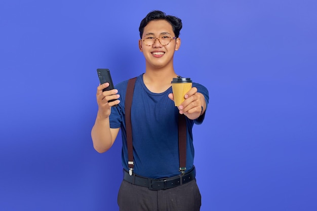 Smiling young man holding smartphone and cup of coffee on purple background
