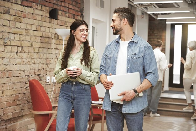 Smiling young man holds laptop talks with female colleague with coffee in office Coworking concept
