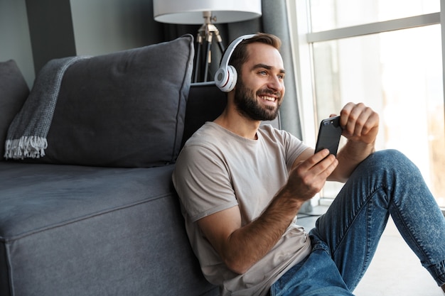 自宅で室内でヘッドフォンで音楽を聴いている笑顔の若い男が床に座って携帯電話でおしゃべりしています。