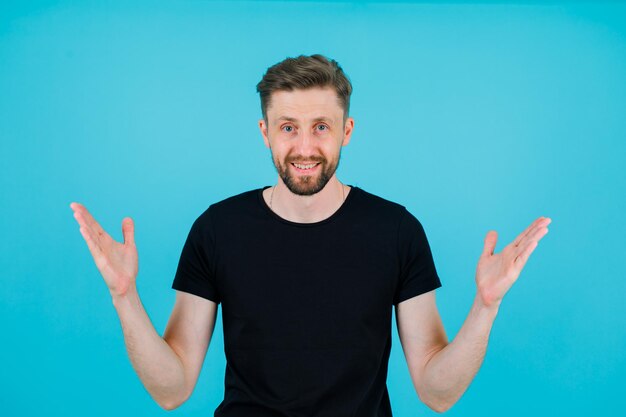 Smiling young man is looking at camera by opening wide hands on blue background