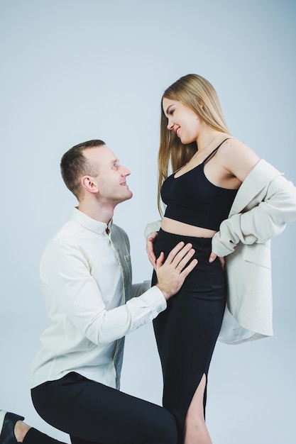 Smiling young man kneeling with his pregnant wife and hugging her belly isolated on white background Stylish young people A man in a shirt and a woman in a black skirt and jacket