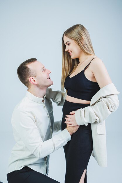 Smiling young man kneeling with his pregnant wife and hugging her belly isolated on white background Stylish young people A man in a shirt and a woman in a black skirt and jacket