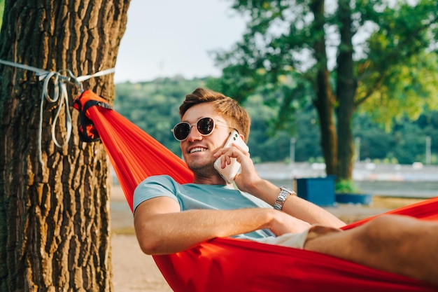 笑顔の若い男が公園で日没時にハンモックに横たわって、笑顔で電話をかけます