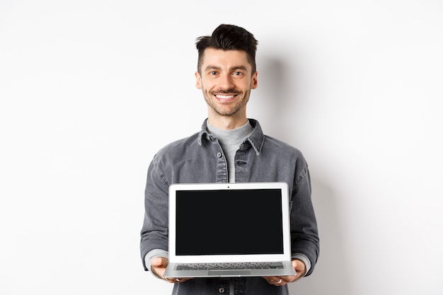 Smiling young man showing empty laptop screen, showing online deal, standing on white background.