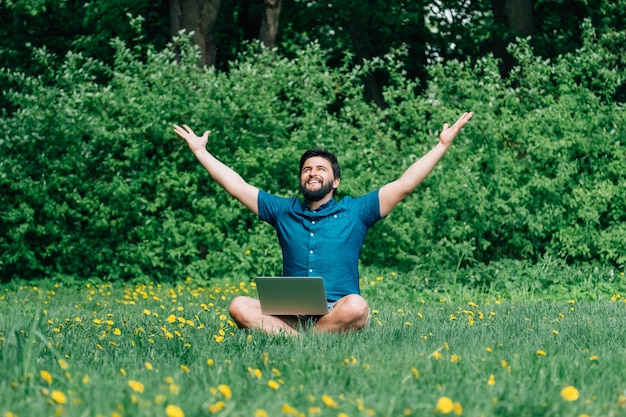 Photo smiling young man sitting on green grass with raised his hands up and with laptop on his crossed legs