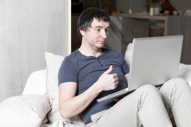 Улыбающийся молодой человек, сидя на диване с ноутбуком. Удаленная работа во время пандемии коронавируса.
