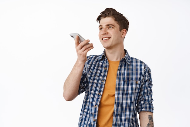 smiling young man talking on speakerphone, record voice message or translate speach with mobile phone app, standing casual on white.