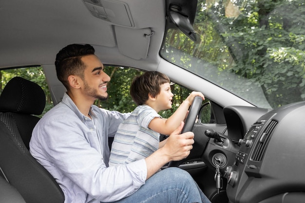 夏の男の子に車を運転するように小さな子供を教えている笑顔の若い男はハンドルを握って見て