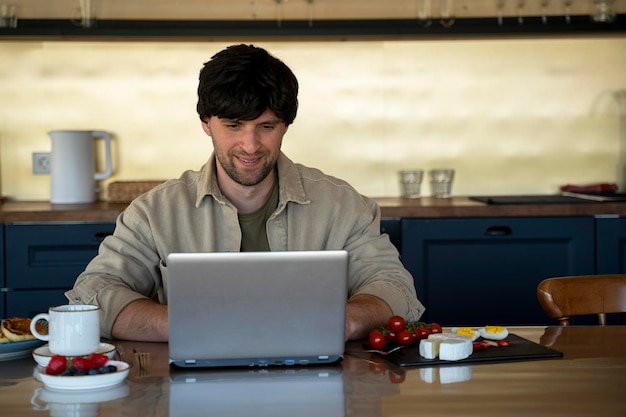 Smiling young man uses a laptop and has breakfast in the kitchen