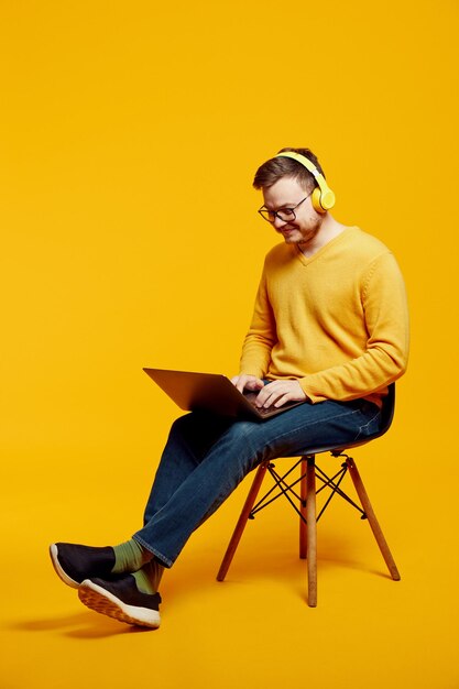 Foto giovane sorridente che usa un portatile e ascolta musica con le cuffie gialle mentre è seduto sulla sedia