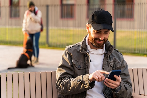 거리에서 현대적인 스마트폰을 사용하여 웃고 있는 청년. 벤치에 앉아 메시지를 입력하는 동안 화면을 가리키는 예쁜 갈색 머리 남자. 현대 생활 개념
