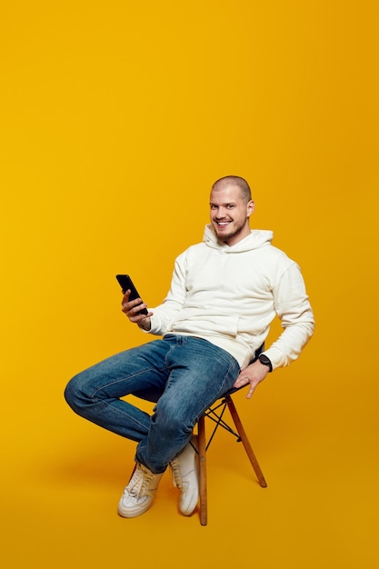 Smiling young man using smartphone while sitting on chair isolated on yellow background