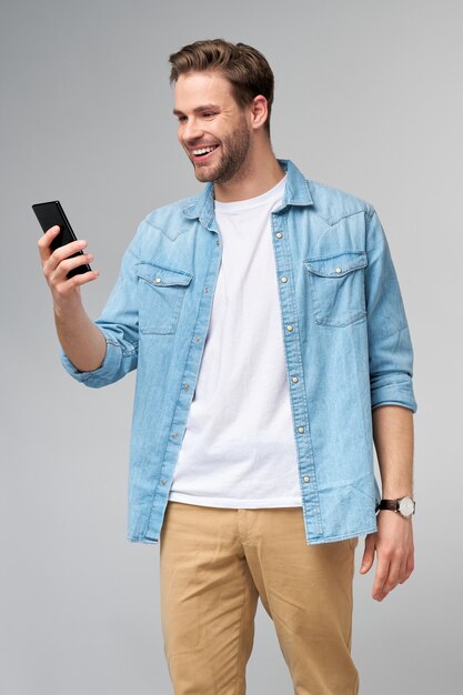 Smiling young man wearing jeans shirt taking selfie photo on smartphone or making video call standing  