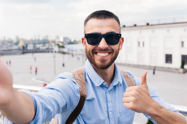 Occhiali da sole da portare sorridenti del giovane che prendono selfie e che mostrano pollice sul gesto