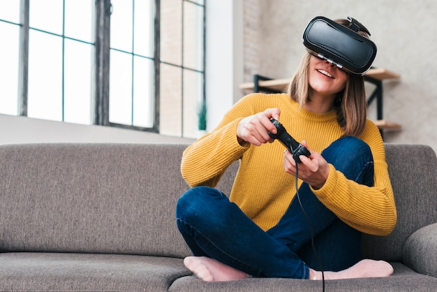 Smiling young man wearing virtual reality glasses sitting on sofa playing video game with joysticks