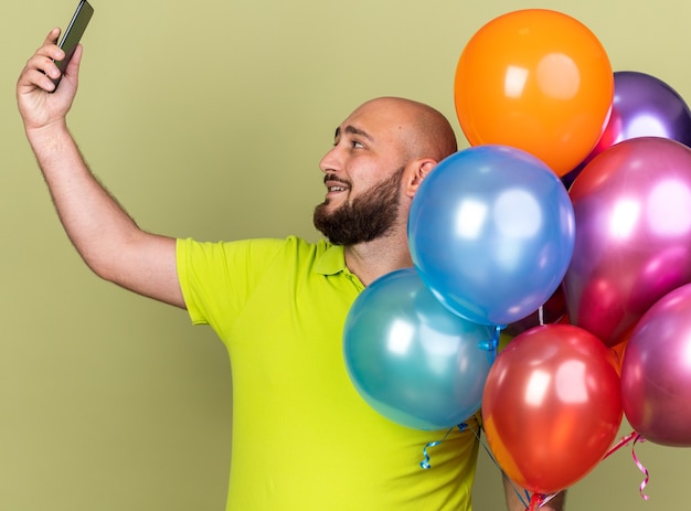 Smiling young man wearing yellow t-shirt holding balloons take selfie isolated on olive green wall
