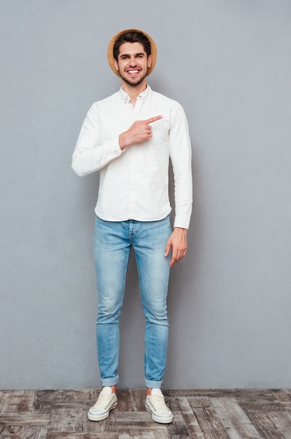 Smiling young man in white shirt and hat standing and pointing away