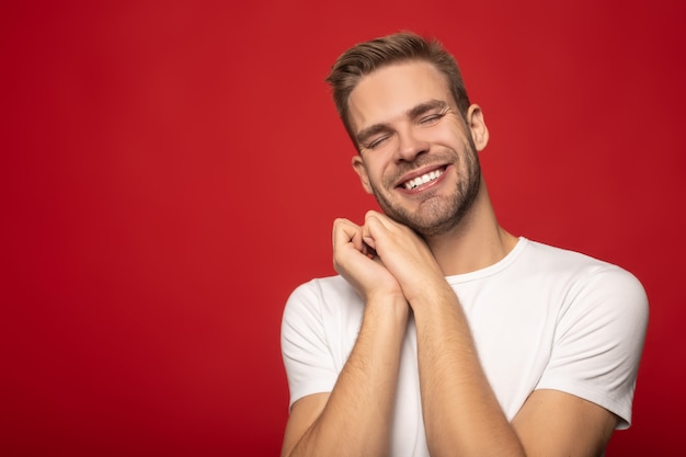 smiling young man with closed eyes and hands near face isolated on red