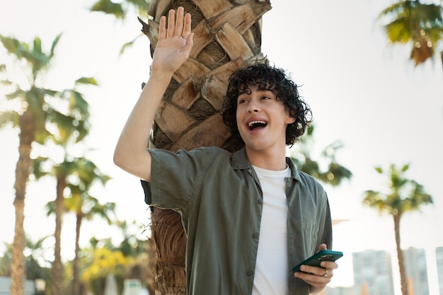 Smiling young man with curly hair waving to somebody while standing with smartphone