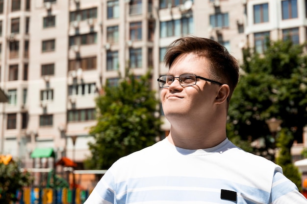 A smiling young man with Down syndrome in glasses poses against the background of the city in the summer