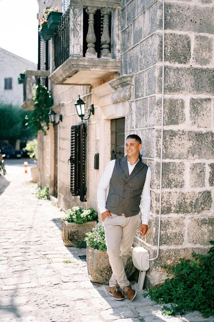 Photo smiling young man with his hand in his trouser pocket is leaning against the wall of an old stone