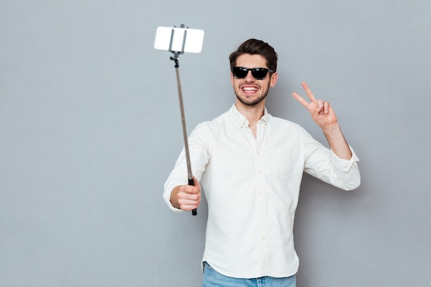 Giovane sorridente con occhiali da sole che scatta foto con smartphone e bastone selfie isolato