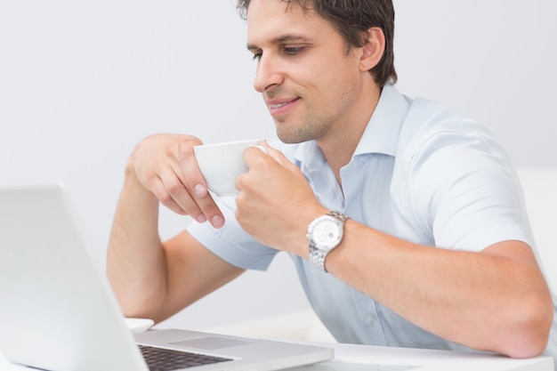 Улыбающийся молодой человек с чашкой, используя ноутбук дома
