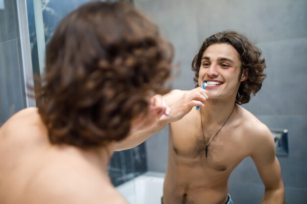 Smiling young man with toothbrush cleaning teeth and looking to the mirror at bathroom