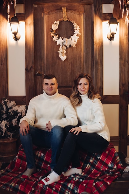 Smiling young man and woman holding hands while sitting on the porch of the house with Christmas decoration on it.