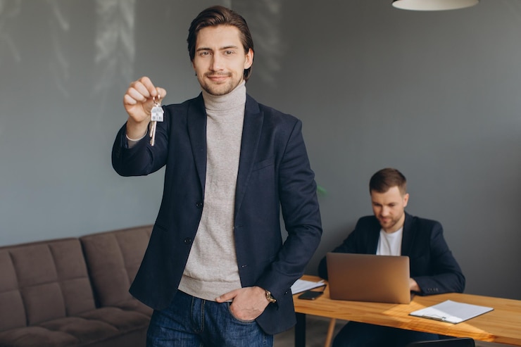 Photo smiling young modern man in a suit holds the keys to the bought apartment on the background of the realtor and office