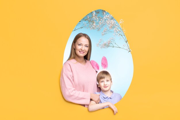 Smiling young mother and cute son in bunny ears headband posing in egg-shaped frame for family Easter card