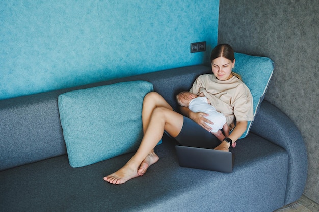 A smiling young mother is breastfeeding her baby and working at home with her baby on the sofa while writing an email Breastfeeding the baby A young woman works at home