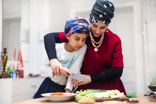 Smiling young mother and pretty little girl in aprons cut fresh salad on wooden board with sharp knife at table in large comfortable kitchen at home