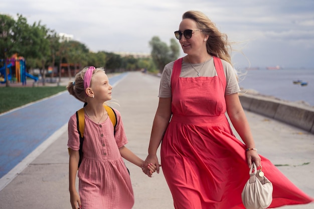 Smiling young mother and schoolgirl walk along embankment smiling and holding hands