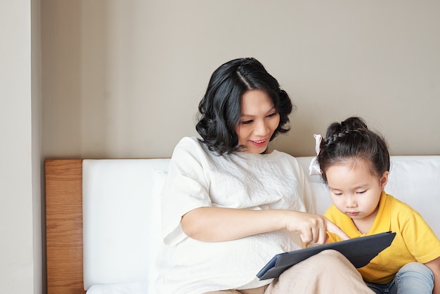 그녀의 어린 딸에게 태블릿 컴퓨터에 교육 응용 프로그램을 보여주는 웃는 젊은 어머니