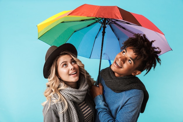 Улыбающаяся молодая многорасовая пара в шарфах стоит с зонтиком, изолированным над синей стеной