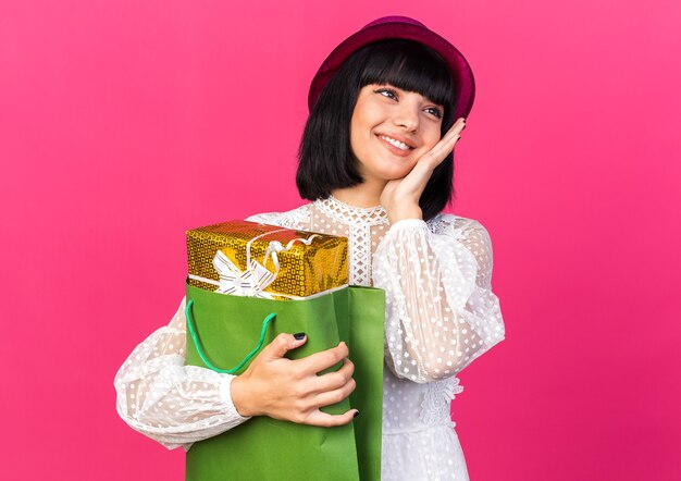 Smiling young party woman wearing party hat holding gift package in paper bag keeping hand on face looking at side isolated on pink wall